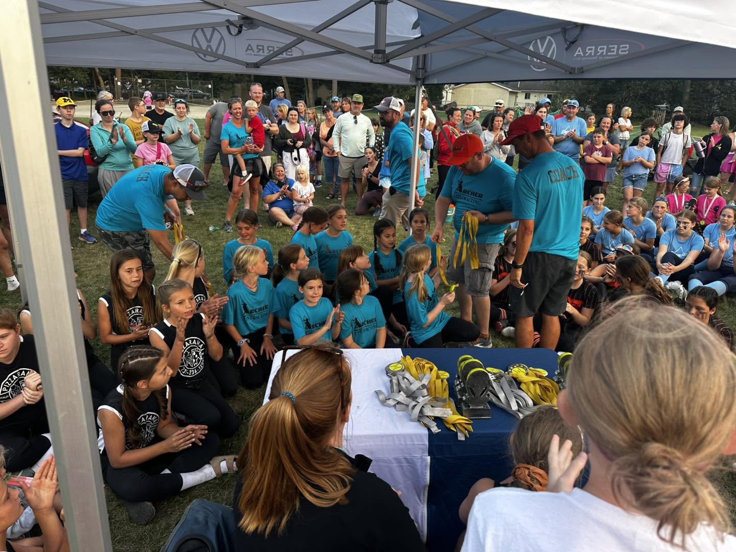 Group of people, including children and adults, gathered under a tent with a table and equipment.